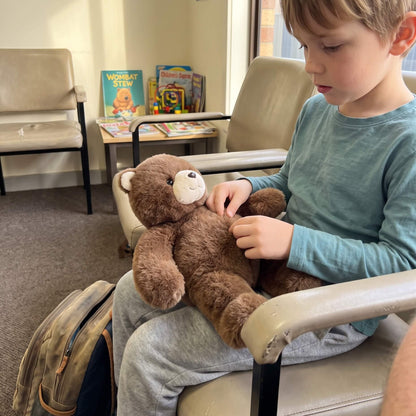 A young boy in a teal long-sleeve shirt and gray sweatpants sits in a beige office chair, holding a brown plush teddy bear. The setting appears to be a clinic or waiting room with other chairs, a small table with books, and posters on the wall.