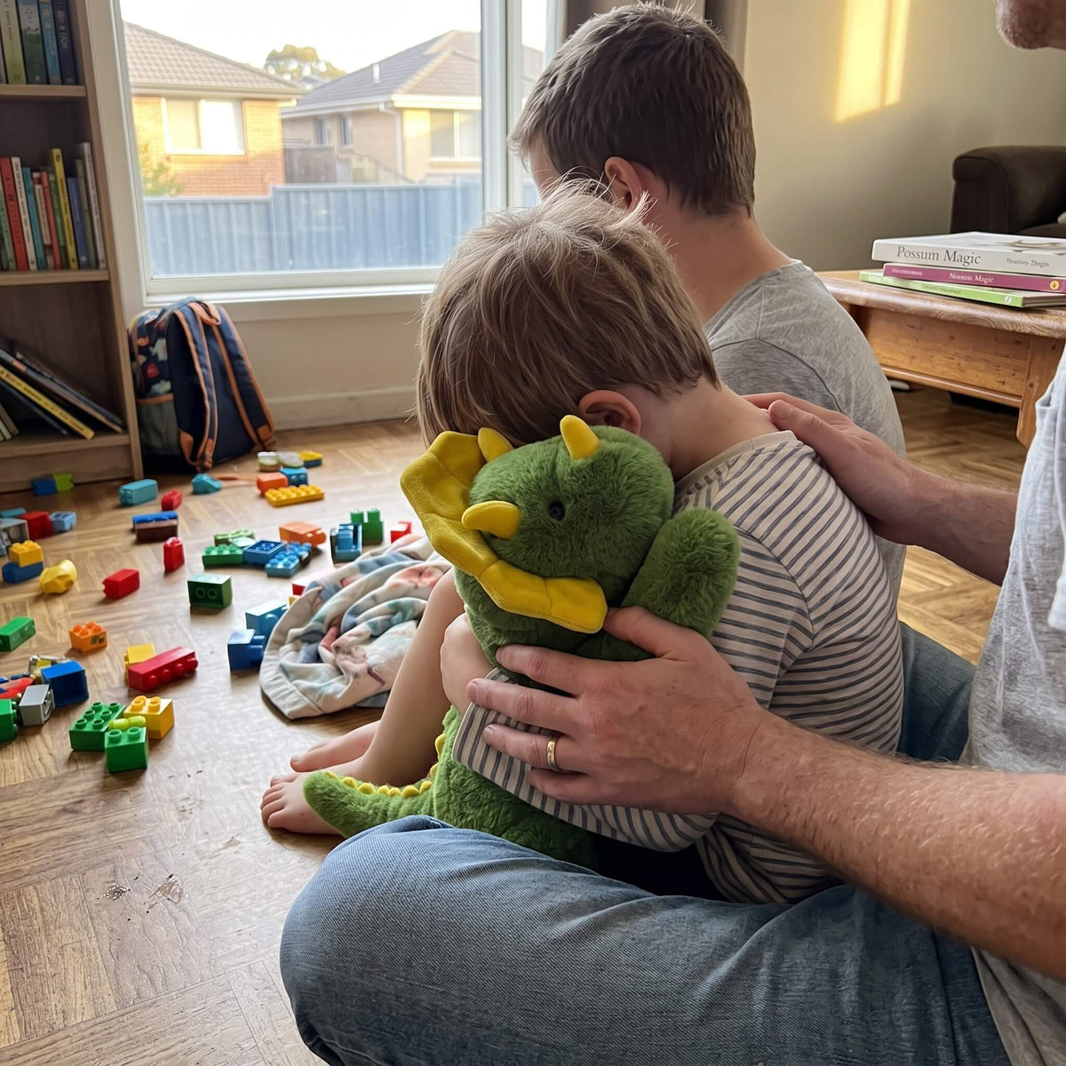 A man and a young boy sit on a wooden floor in a living room, with the man holding a green plush dragon toy with yellow wings and horns. Colorful toy blocks are scattered on the floor nearby.